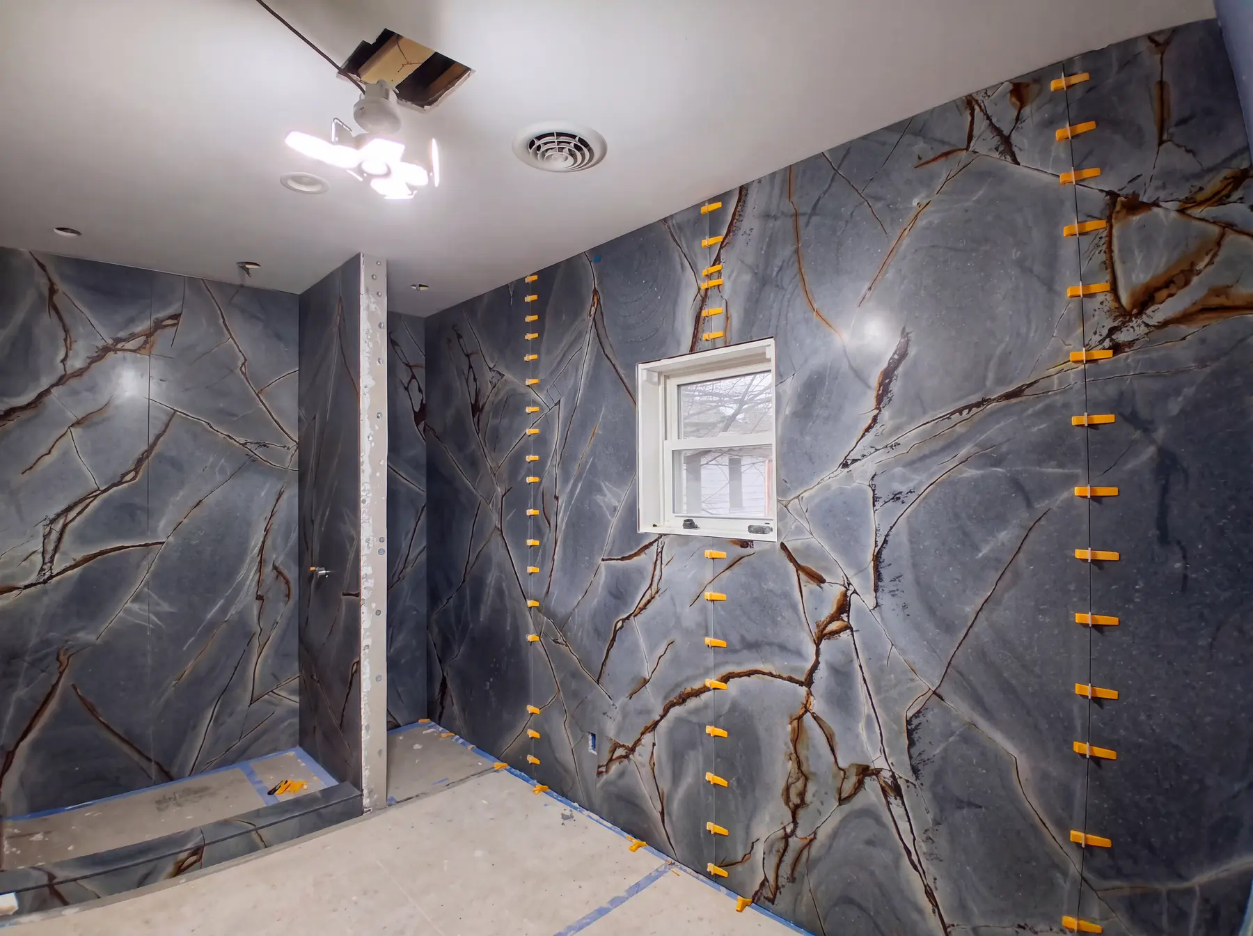A bathroom remodel in progress: large-format marble-effect slab panels set on the walls with levelling clips still in place beside a formed shower niche.