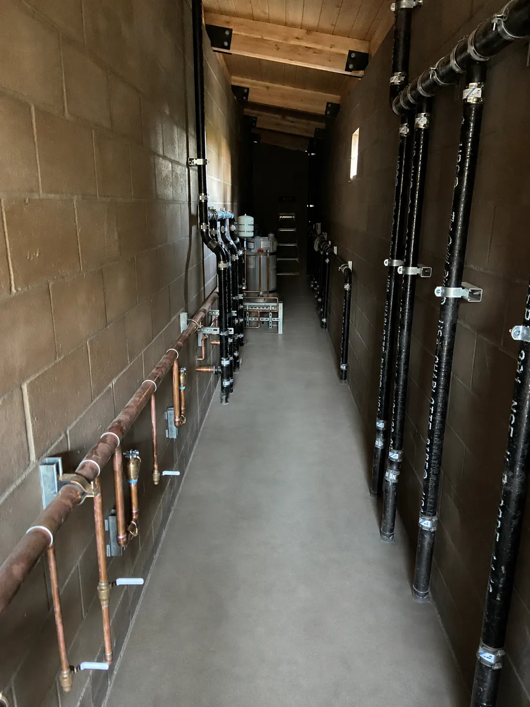 A commercial plumbing chase: a copper valve manifold running down one block wall, black grooved-steel risers down the other, and a water heater at the far end of the run.