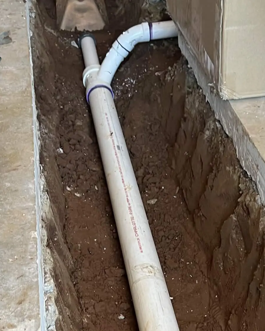 A white PVC supply line running the length of a narrow trench cut through a concrete floor, from its fitting at the top down to the foot of the frame.