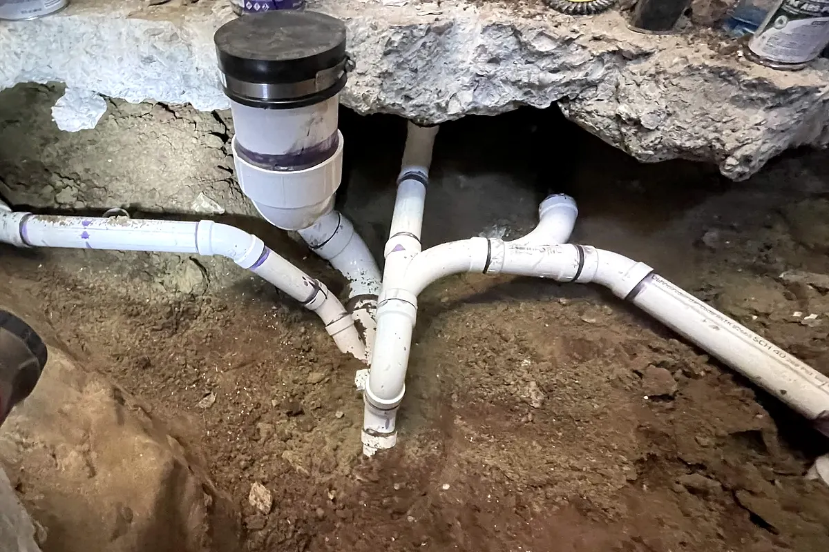 White PVC drain, waste and vent branches joined into a wye beneath a broken-out concrete slab in an open excavation.
